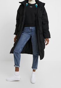 Lange zwarte donsjas, zwart sweatshirt met groene accenten, blauwe spijkerbroek en witte sneakers. Gladde textuur en losse pasvorm.