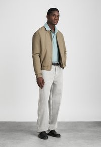 Brun bomberjakke over en lyseblå polo, sammen med lysegrå vide bukser og svarte sko. Enkel bakgrunn med nøytrale toner.