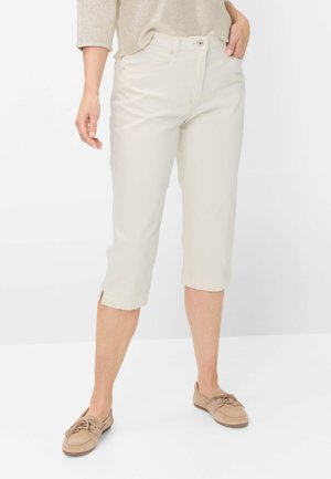 Kvinde iført beige cropped bukser med små sideslids og beige loafers, stående mod en hvid baggrund.