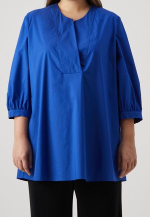 Femme portant un chemisier ample bleu vif à manches trois quarts et un décolleté carré profond, associé à un pantalon noir.