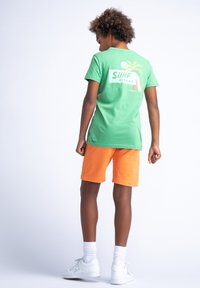 Zelené bavlněné tričko s grafikou palmy a nápisem "SURF RIDERS" na zádech, doplněné oranžovými šortkami. Bílé tenisky a ponožky do kotníků.