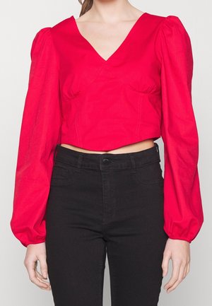 Vrouw die een rood cropped blouse met V-hals draagt, met lange pofmouwen en een high-waisted zwarte spijkerbroek, weergegeven van schouders tot heupen.