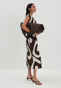 Braun-weißer abstrakt gemusterter Kleid, glattes Material, knielang. Schwarze geflochtene Tasche mit großer Öffnung, kurze glänzende Griffe, minimalistische Sandalen.