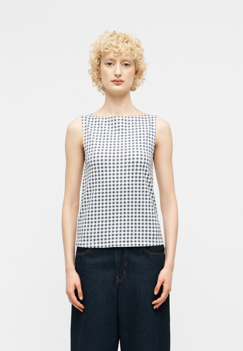 Femme blonde aux cheveux bouclés portant un haut sans manches à carreaux bleu marine et blanc ainsi qu'un jean à jambes larges foncé, debout devant un fond uni.