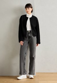 Giacca nera con texture, con zip frontale, indossata sopra un dolcevita bianco e abbinata a jeans grigi a vita alta e scarpe bianche.
