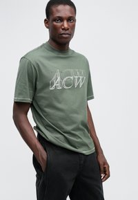 Tricou din bumbac verde măsliniu cu imprimeu alb, având literele "ACW". Mânici scurte și decolteu rotund, purtat cu pantaloni negri.