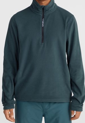 Mężczyzna noszący ciemnoturkusową bluzę polarową z suwakiem do połowy z przodu oraz logo na lewym rękawie i zamku.