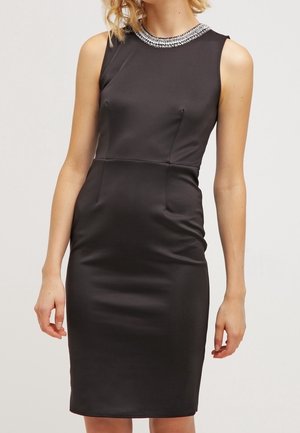 Hihaton musta vartalonmyötäinen mekko polvenpituinen helma ja koristeltu korukivet kaulus, jonka käyttää henkilö, jolla on löysät vaaleat hiukset.