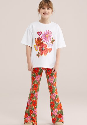 Niña sonriente, con camisa blanca de gran tamaño con estampado colorido de flores y corazones, y pantalones acampanados vibrantes con diseño floral.