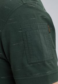 Tela verde texturizada con un pequeño bolsillo de parche en el hombro. Presenta un patrón sutil con vetas de verde más claro.