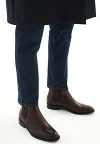 Botine Chelsea din piele maro, cu finisaj neted, laterale elastice și vârf rotunjit, purtate cu blugi slim-fit de culoare albastru închis.