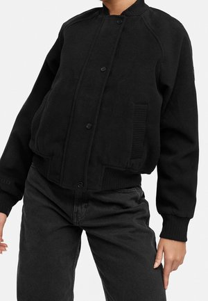 Personne portant une veste bomber noire boutonnée avec des poignets, un col et une ceinture côtelés, associée à un pantalon noir taille haute.