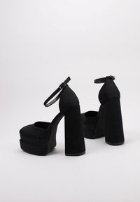 Zapatos de plataforma negros de tacón alto con tacones gruesos, puntera redondeada y correas en el tobillo. Hechos de un material suave con una textura lisa.