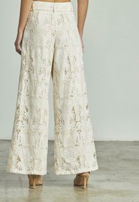 Pantaloni a gamba larga realizzati in pizzo elaborato, di colore crema con motivi ricamati. La cintura è aderente e le scarpe con il tacco sono visibili.