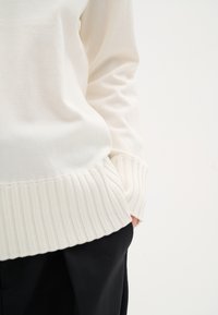 Maglione a maglia off-white con maniche lunghe, caratterizzato da un orlo a coste. Abbinato a pantaloni neri, che mostrano dettagli ai polsini e in vita.