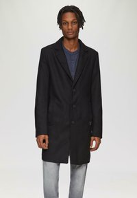 Schwarzer langer Mantel mit Reverskragen, ausgestattet mit vier vorderen Knöpfen und Seitentaschen, kombiniert mit einem blauen Hemd mit Knöpfen und hellen Jeans.