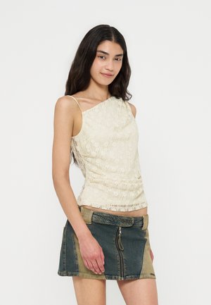 Jeune femme aux longs cheveux foncés portant un haut en dentelle crème à une épaule et une mini-jupe en jean bicolore avec fermeture éclair à l'avant.