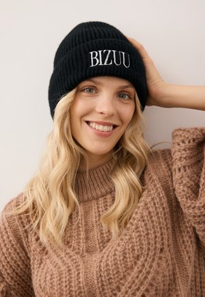 Zwarte geribde beanie met "BIZUU" borduring. Zachte textuur, aansluitend ontwerp, gedragen boven een comfortabele bruine gebreide trui met patroon.