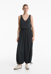 Črn brezrokavnik z V-izrezom, nabranim pasom in ohlapnimi, koničastimi hlačami. Ima teksturirano tkanino in vezalke na robovih. Nosi se z bež športnimi copati.