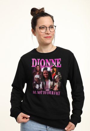 Frau mit Brille und Dutt, die einen schwarzen Pullover trägt, der mehrere Bilder einer Frau und den Text "DIONNE" und "NEIN, NICHT INS GESICHT." zeigt.
