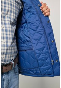 Wnętrze pikowanej niebieskiej kurtki o gładkiej fakturze, z zamykaną na zamek kieszenią i zatrzaskami wzdłuż krawędzi. Noszona na koszuli w kratę.
