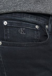 Jeans de denim negro con un sutil brillo, que cuentan con un pequeño logo de CK en el bolsillo, detalles en plata y un diseño clásico de cinco bolsillos.