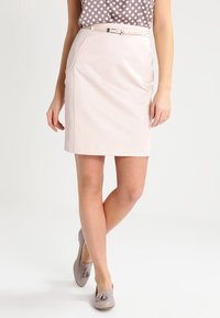 Helles rosa Bleistiftrock mit Seitentaschen und einem sanften Schimmer, in schmaler Passform und mit einem passenden schmalen Gürtel. Kombiniert mit grauen Quasten-Mokassins.