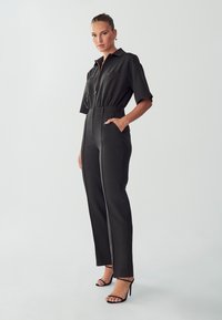 Zwarte jumpsuit met een kraagnek, korte mouwen, ritssluiting, zijzakken en taps toelopende broekspijpen met een verticale naaddetail.