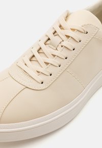 Beige sneakers with a smooth leather upper, round toe, and textured lace area. Features matching flat laces and a white rubber sole.