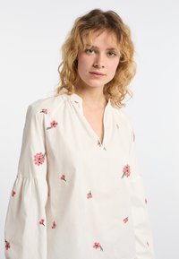 Weißes Baumwollbluse mit V-Ausschnitt und langen Puffärmeln, die mit rosa floralem Stickerei übersät ist.