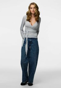 Sivi ovijalni vrh z dolgimi rokavi, z globokim V-izrezom in pasom za zavezovanje v pasu. Kombiniran z modrimi kavbojkami širokih hlačnic.