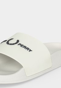 Sandale blanche à semelle plate avec une large sangle présentant un logo de couronne de laurier noire et le texte "PERRY" en haut.