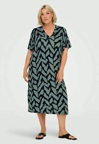 Ξανθιά γυναίκα που στέκεται σε ένα χαλαρό, μήκους γόνατος φόρεμα με V λαιμόκοψη, με μαύρα και γκρι αφηρημένα σχέδια σε ζικ ζακ και μαύρες σαγιονάρες.