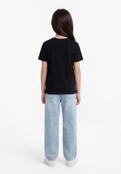 Bambino con lunghi capelli scuri che indossa una t-shirt nera, jeans a gamba larga azzurri chiari e sneakers bianche, in piedi con le spalle rivolte verso l'obiettivo su sfondo bianco.