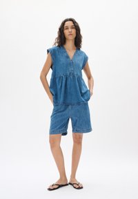 Denim tērps, kas ietver brīvu, īsām piedurknēm, blūzi ar V-veida kakla izgriezumu un savilktu jostu, apvienots ar ceļu garuma džinsu šortiem. Zila krāsa.