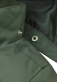 Olijfgroen stof met een gestructureerde afwerking, heeft een drukknoopsluiting en zichtbare stiksels. Opvallende hardware omvat metalen drukknopen.