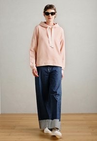 Felpa con cappuccio rosa chiaro dotata di lacci, abbinata a jeans a gamba larga blu scuro con orlo risvoltato, indossata con sneakers bianche.