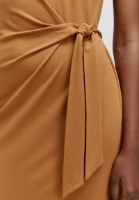 Le tissu de la robe marron présente un détail de taille nouée avec un design fluide. Texture lisse avec un subtil éclat, rehaussé par une longue ceinture.