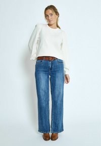 Blusa bianca con maniche a palloncino, abbinata a jeans larghi blu e una cintura marrone con fibbia rotonda. Scarpe con tacco marroni completano l'outfit.