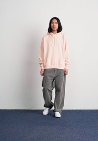 Ροζ φούτερ με κορδόνια, συνδυασμένο με χαλαρές γκρίζες ριγέ παντελόνες και λευκά αθλητικά παπούτσια, σε απλό λευκό φόντο.