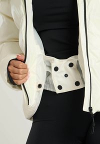 Witte pufferjas met een glanzende afwerking; uitgerust met een zwarte rits, drukknoopjes en een binnenzak. Zwarte basislaag zichtbaar eronder.