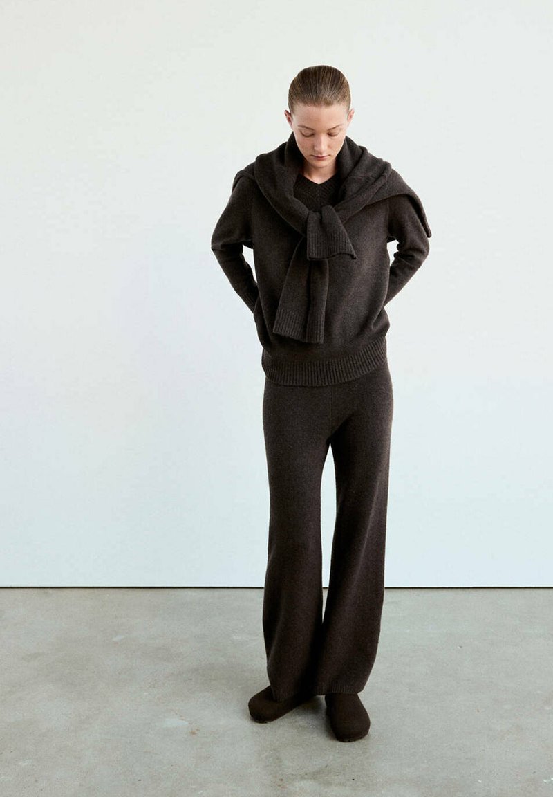 Femme portant un pull en maille marron foncé et un pantalon, avec le pull posé sur les épaules, debout sur un sol gris contre un mur blanc.