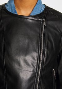 Giacca di pelle nera con colletto strutturato e zip asimmetrica. Texture liscia e design fitted. Jeans visibili sotto.