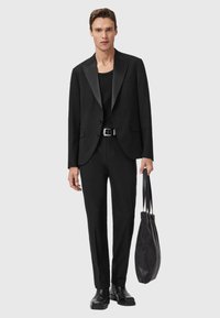 Uomo con blazer nero, camicia nera, pantaloni neri e stivali neri, che tiene una borsa a mano nera texturizzata su uno sfondo chiaro uniforme.