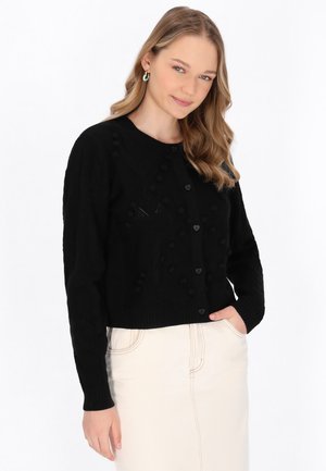 Cardigan corto nero con lavorazione a maglia testurizzata, bottoni a forma di cuore e dettagli a pompon sulla parte anteriore, abbinato a una gonna chiara.