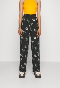 Czarne kwieciste spodnie z zielonymi liśćmi i różowymi kwiatami. Wysoki stan, szerokie nogawki, zestawione z białymi i czarnymi sandałami na platformie.