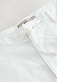 Valged linased lühikesed püksid nööriga seotud sulgemiskohaga, millel on sile tekstuur ja puhas disain. Silt näha vöökohal seespool.