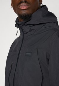 Black weather-resistant jacket featuring a hood, zip front, and chest pocket with a logo patch. Smooth texture with a matte finish.