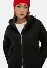 Zwarte fleecetrui met rits en capuchon, compleet met twee voorzakken en een zilveren ritsaccent. Afgemaakt met een rode gebreide beanie.