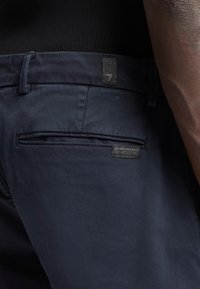 Pantalons bleu marine au toucher lisse, présentant une poche arrière et une étiquette en tissu près de la taille. Design minimal et forme épurée.